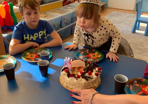 6 urodziny Nikoli. Na stoliku stoi tort. Nikola pochylona nad tortem dmucha na świeczki. Obok Nikoli siedzi chłopiec w niebieskiej koszulce.